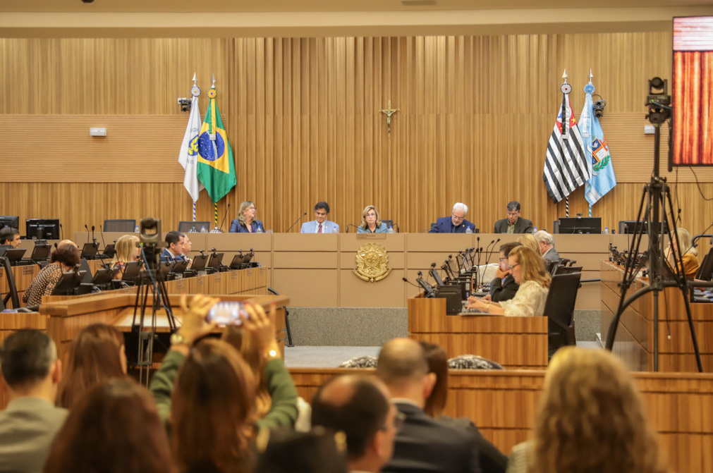 plenário com público em primeiro plano de costas e mesa alta composta pelas autoridades