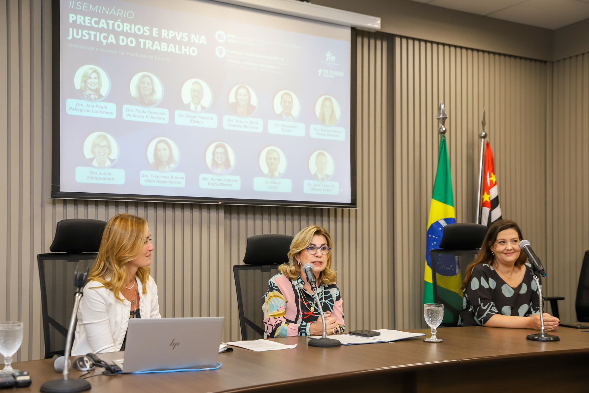 Mesa de encerramento com a Dra. Ana Paula no centro, tendo a Dra. Daniela de seu lado direito e a servidora Samira, do lado esquerdo. Ao fundo, o telão temático e bandeiras
