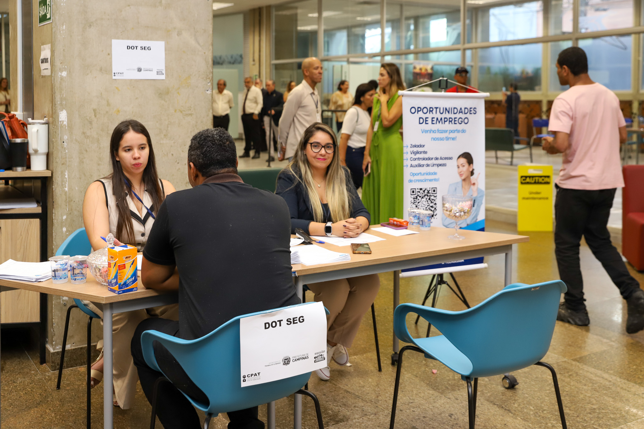 imagem de trabalhador negro sendo entrevistado por uma analista de recrutamento, ao lado de outra, que olha para a câmera. Todos sentados à mesa. Ao lado, um banner com os dizeres: Oportunidade de Emprego