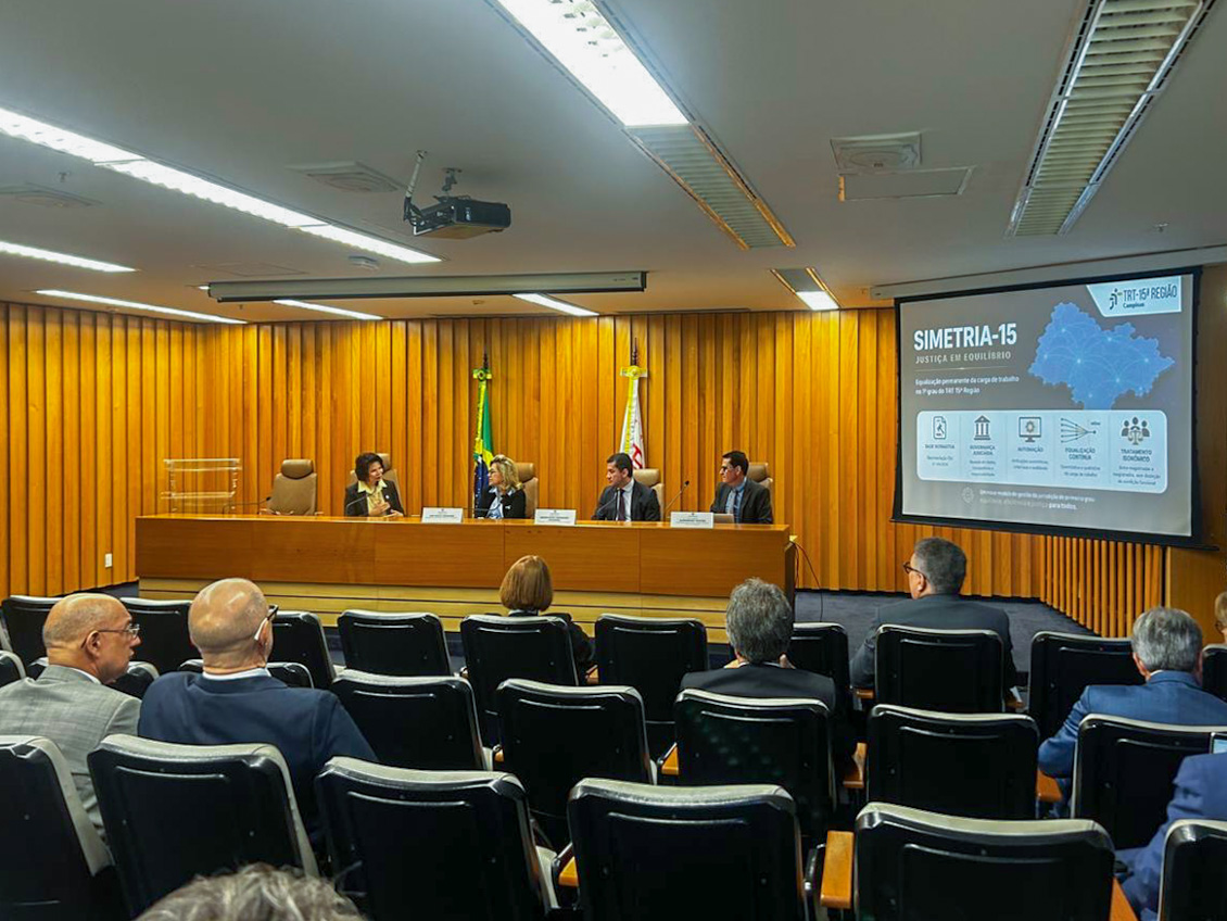À frente da sala, quatro autoridades estão sentadas atrás de uma mesa de madeira, diante das bandeiras do Brasil e do Estado de São Paulo. À direita, um telão exibe slide do projeto apresentado pelo TRT-15. Na plateia, participantes acompanham a exposição sentados em cadeiras pretas dispostas em fileiras. O ambiente possui iluminação clara e paredes revestidas em madeira.