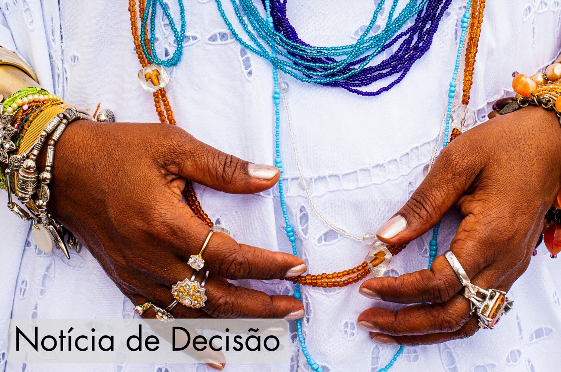 Imagem em close das mãos de uma pessoa de pele negra posicionadas à frente do corpo, segurando delicadamente um colar de contas em tons de âmbar. A pessoa veste uma roupa branca rendada e usa diversos colares de miçangas sobrepostos, em tons de azul, turquesa, roxo, branco e âmbar. Nos dois pulsos há várias pulseiras metálicas e de miçangas coloridas, e em ambas as mãos há diversos anéis prateados e dourados. O fundo é preenchido pela própria vestimenta branca, destacando as cores vibrantes dos acessórios. 