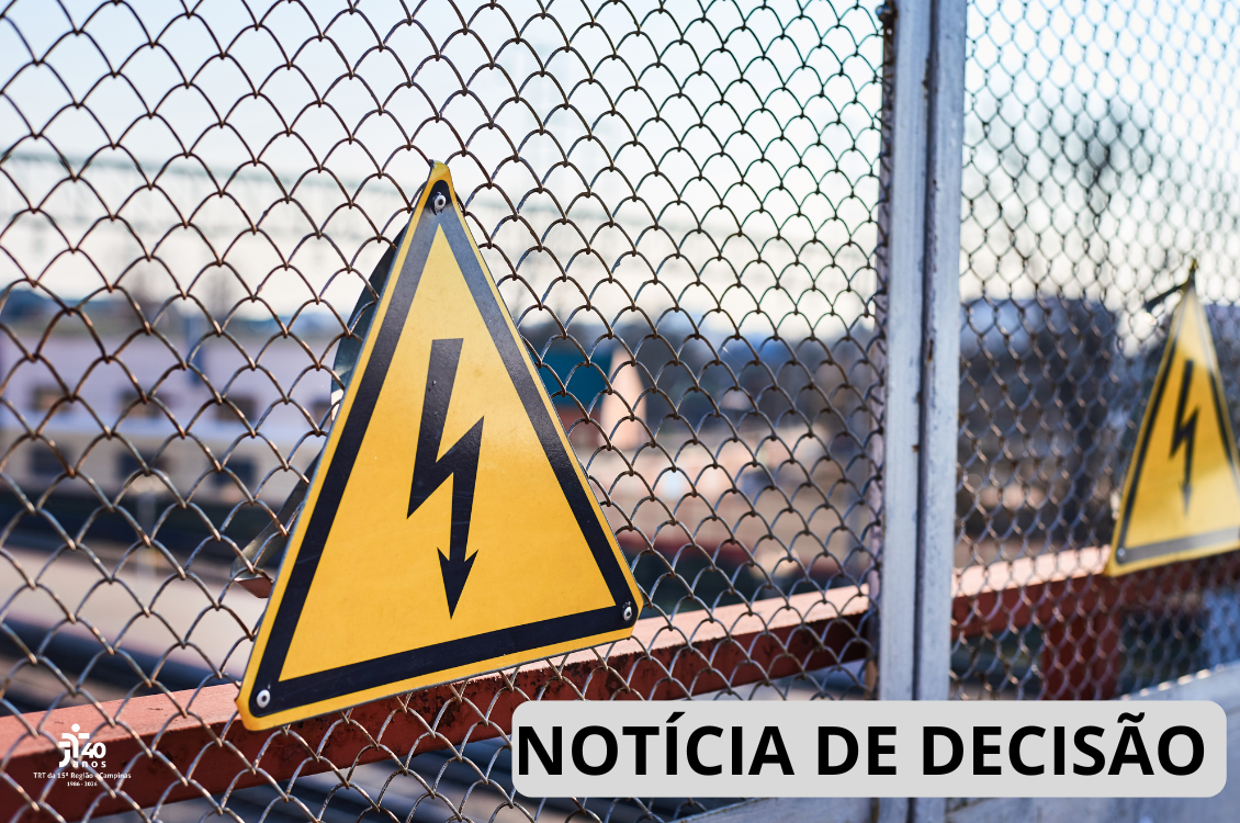 : Ilustração mostra, em primeiro plano, uma placa triangular amarela de advertência para risco de choque elétrico, com borda preta e símbolo de um raio, afixada em uma cerca de tela metálica. Ao fundo, desfocados, aparecem estruturas e equipamentos de uma área industrial ou subestação elétrica, além de uma segunda placa de advertência semelhante. No canto inferior direito, há a inscrição