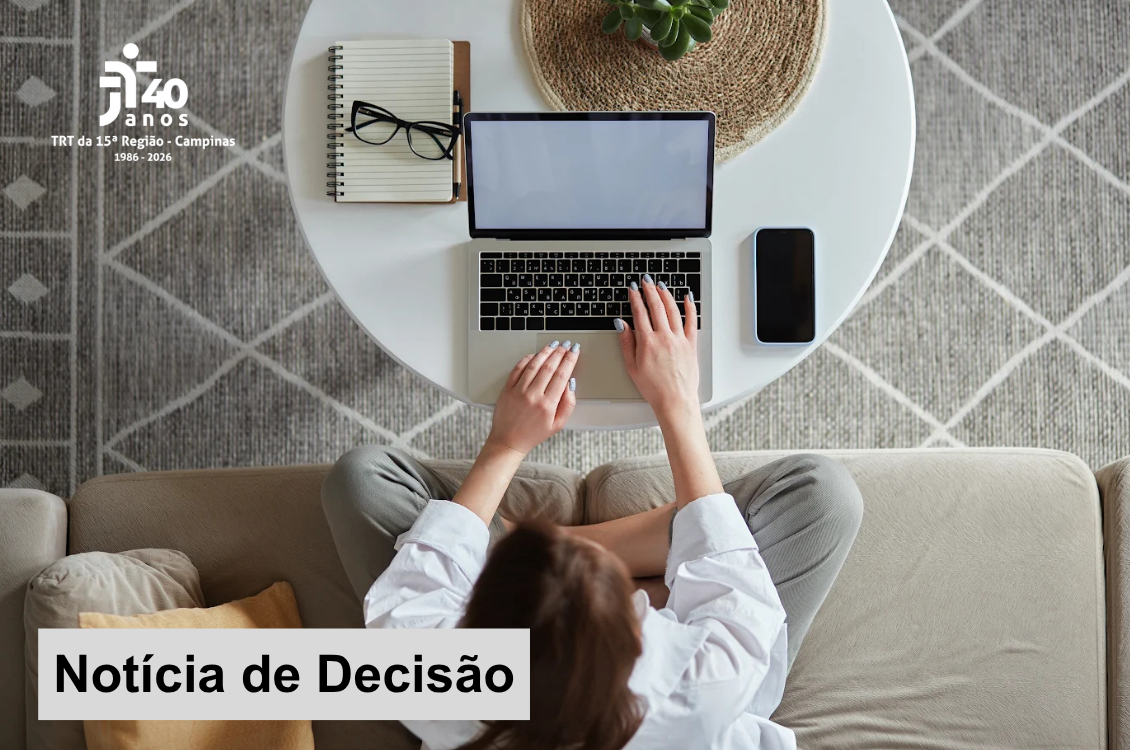 Imagem em formato horizontal, vista de cima. Uma pessoa sentada em um sofá bege utiliza um notebook apoiado sobre uma mesa redonda branca. Ao lado do computador há um celular, um caderno com óculos e, na parte superior, um vaso com uma pequena planta. A pessoa veste camisa branca e calça cinza. Sobre a imagem, no canto superior esquerdo, aparece o logotipo dos 40 anos do TRT da 15ª Região – Campinas, com o período “1986–2026”. Na parte inferior esquerda, há uma faixa branca com a inscrição em letras pretas: