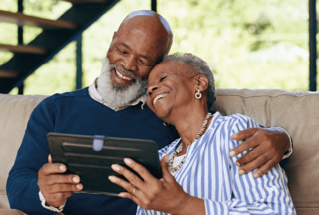 Foto de dois idosos abraçados sentados. Eles seguram um tablet e sorriem.