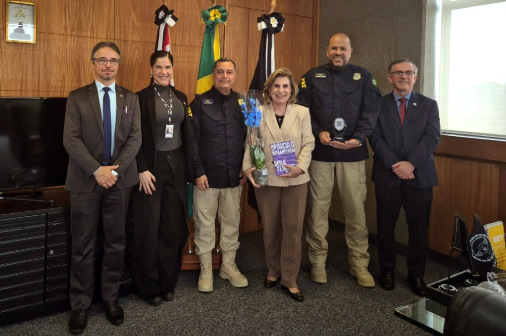 foto posada com Dra. Ana Paula, segurando um livro e uma orquídea, com o assessor Cleber, Dr. João e três representantes da PRF, sendo uma mulher