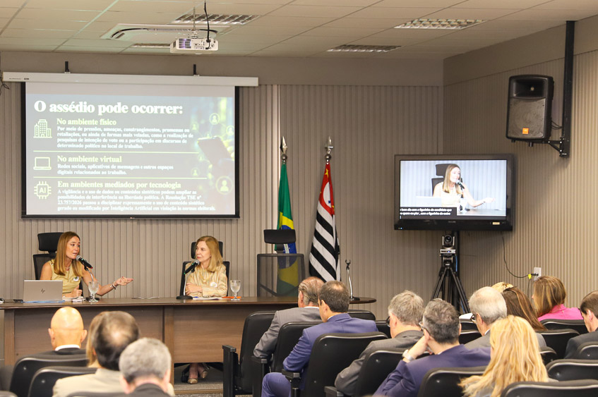Dra. Daniela e Dra. Antonia na mesa de trabalhos com público em primeiro plano e telão ao fundo
